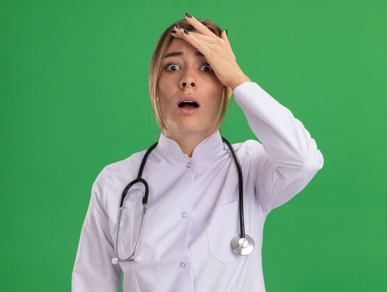 害怕年轻的女医生被吓坏了 穿着医用长袍 手放在额上 隔着绿墙长袍听诊器手
