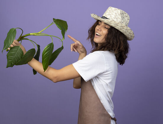 成人年轻的女园丁面带微笑 穿着制服 戴着园艺帽 指着植物紫色园艺微笑