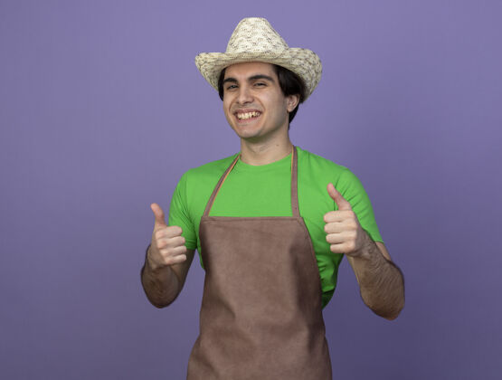 表演微笑着的年轻男园丁穿着制服 戴着园艺帽 竖起大拇指人紫色花园