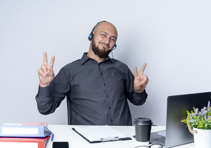 年轻高兴的年轻秃头呼叫中心男子戴着耳机坐在办公桌旁与工作工具做和平的迹象隔离在白色的背景工作中心电话