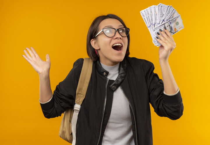 钱印象深刻的年轻女学生戴着眼镜 背着书包抱着钱 孤立地看着橘子年轻橘子包