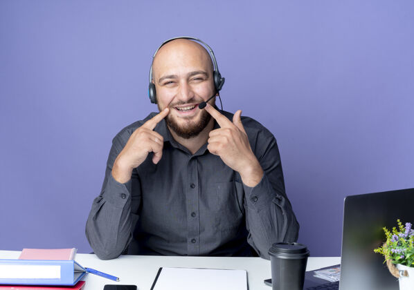 相机年轻秃顶的呼叫中心男子戴着耳机坐在办公桌旁 手里拿着工作工具 手指放在嘴边 假装微笑 看着紫色背景上孤立的摄像头手指穿工作