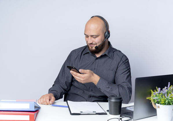 中心高兴的年轻秃头呼叫中心男子戴着耳机坐在办公桌旁 拿着工作工具 看着白色背景上孤立的手机工具年轻通话