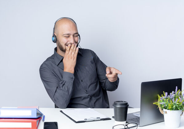 电话戴着耳机的年轻秃头呼叫中心男子坐在办公桌旁 手里拿着工作工具 一边看着笔记本电脑 一边用手捂着嘴 隔离在白色背景上工作中心秃头