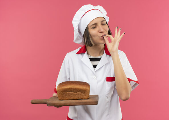 面包身着厨师制服的年轻女厨师手拿着放着面包的砧板 做着可口的手势 闭着眼睛 在粉红色的背景上与世隔绝请好吃切