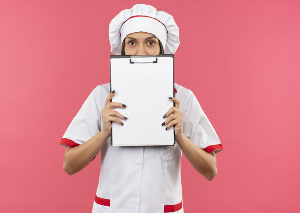 相机年轻的女厨师穿着厨师制服 从剪贴板后面拿着相机看着 背景是粉色的 还有复印空间厨师拿着烹饪