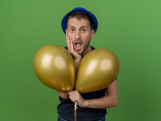 氦戴着蓝色派对帽的帅哥惊呆了 把手放在脸上 拿着氦气球隔离在绿色的墙上 留着复制空间表情党气球