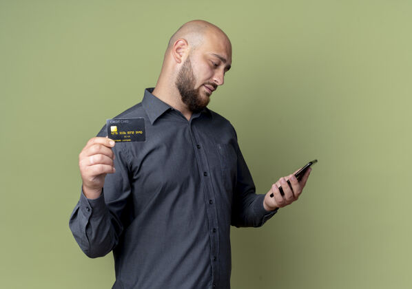 橄榄年轻的秃头呼叫中心男子拿着手机 看着手机和信用卡在橄榄绿的背景与复制空间隔离相机年轻秃头电话