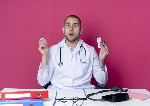长袍年轻的男医生穿着医用长袍 听诊器坐在办公桌旁 手里拿着工作工具 手里拿着医用烧杯 一包包药片和胶囊隔离在粉红色的桌子上穿胶囊印象