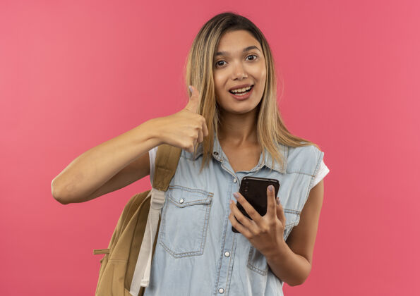 高兴快乐的年轻漂亮的学生女孩戴着背袋拿着手机 在粉红色的地板上竖起大拇指背手机包