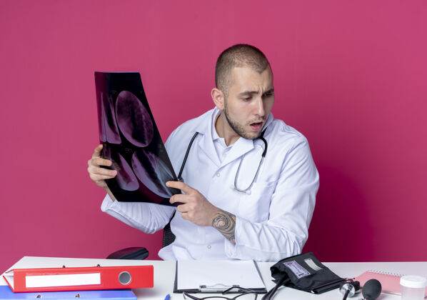 印象年轻的男医生穿着医用长袍 手持听诊器坐在办公桌旁 手里拿着工作工具 手里拿着x光片 看着粉红色的办公桌男性穿着医疗