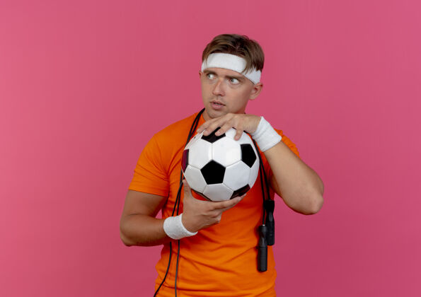 头带印象深刻的年轻英俊的运动男子戴着头带和腕带与跳绳脖子上拿着足球看一边孤立的粉红色印象帅气周围