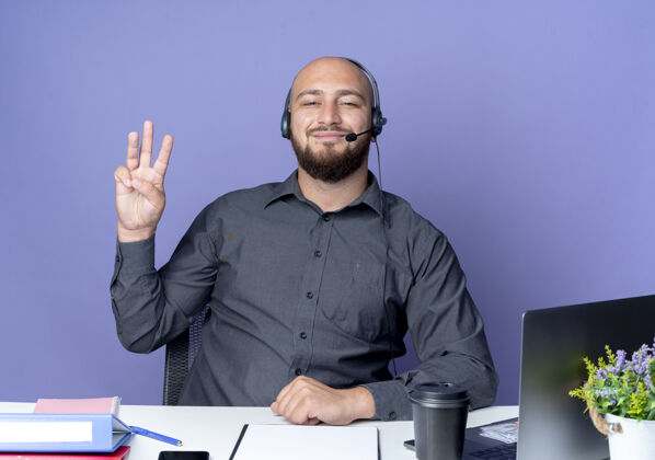 请高兴的年轻秃头呼叫中心男子戴着耳机坐在办公桌上 工作工具显示三个孤立的紫色背景中心秃头年轻