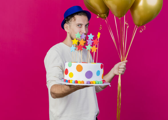 男人高兴的年轻英俊的斯拉夫党家伙戴着党的帽子拿着气球和生日蛋糕与星星伸展向前方看前面的粉红色墙上孤立的复制空间年轻穿着人