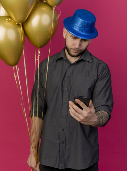 帽子皱眉的年轻帅哥斯拉夫党人戴着党帽手持气球和手机看着手机孤立在粉红色的墙上皱眉气球抱着