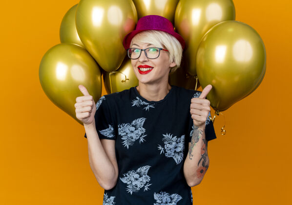 人微笑着的金发派对女郎戴着派对帽戴着眼镜站在气球前看着前面竖起大拇指孤立地站在橙色的墙上衣服穿着眼镜