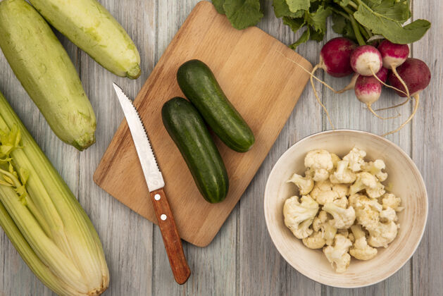 芹菜顶视图新鲜的绿色黄瓜在一个木制的菜板上 刀子上放着菜花芽 碗里放着萝卜 西葫芦和芹菜 背景是灰色的木制花椰菜观点刀
