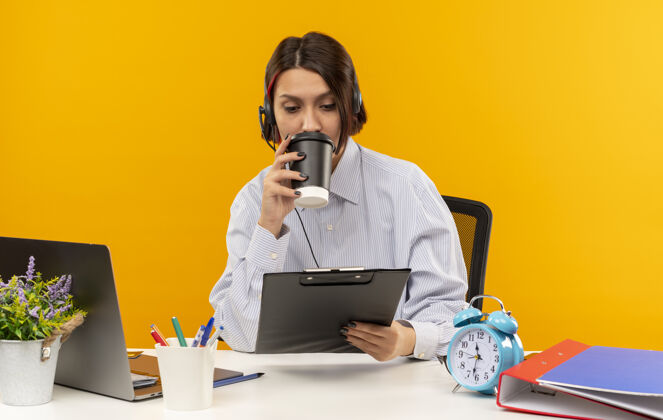 工作年轻的呼叫中心女孩戴着耳机坐在办公桌旁 手里拿着工作工具 拿着塑料咖啡杯喝着咖啡 看着孤立在橙色背景上的剪贴板塑料坐着工具