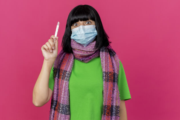 表情印象深刻的年轻生病的女人戴着口罩和围巾拿着温度计看着前面隔离在粉红色墙上的复制空间公民人衣服