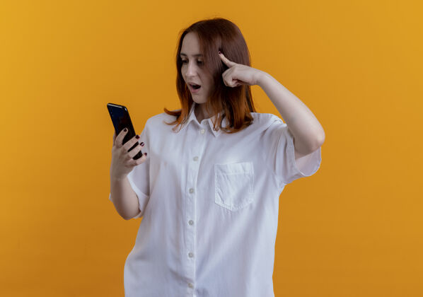 惊喜惊讶的年轻红发女孩拿着手机 看着手机 把手指放在隔离在黄色背景上的头上拿着头电话