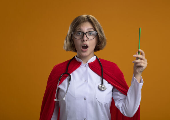 铅笔令人印象深刻的年轻金发女超级英雄在红色斗篷穿着医生制服和眼镜听诊器拿着铅笔看前面孤立的橙色墙壁与复制空间制服金发眼镜
