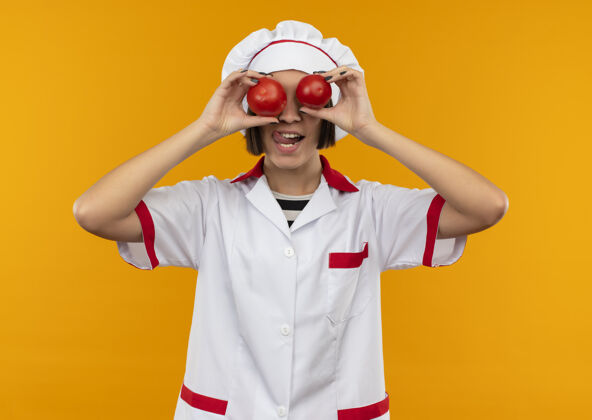 烹饪穿着厨师制服的顽皮年轻女厨师把西红柿放在眼睛上 在橙色背景上露出孤立的舌头眼睛厨师舌头