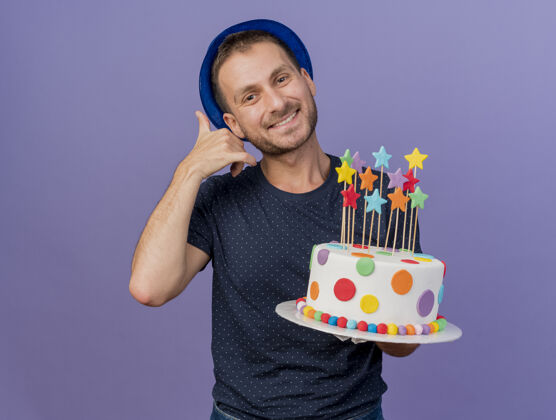 电话面带微笑的帅哥戴着蓝色帽子 手势叫我签名 拿着生日蛋糕孤立地放在紫色的墙上 留着复制空间抱着帅哥人