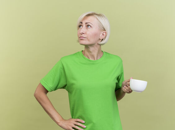 表情体贴的中年金发斯拉夫女人捧着一杯茶 手放在腰上 看着橄榄绿墙上孤立的一面壁板感觉人