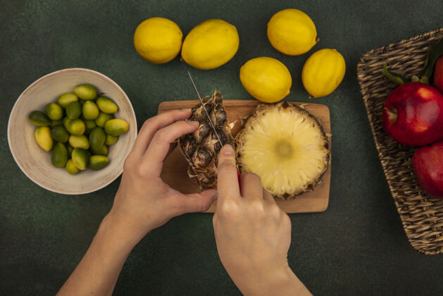 刀俯视图中的女性手切割新鲜菠萝在木制厨房板上用刀与金盏花在一个碗与柠檬隔离在一个绿色的背景女性绿色水果