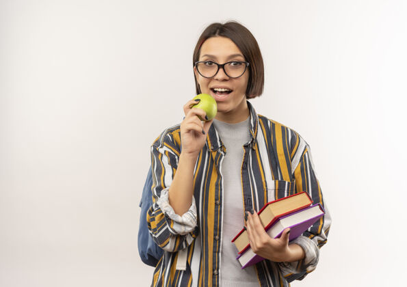 包年轻的女学生戴着眼镜 背着书包 拿着书 准备咬一口苹果 背景是白色的 有复印空间学生准备穿