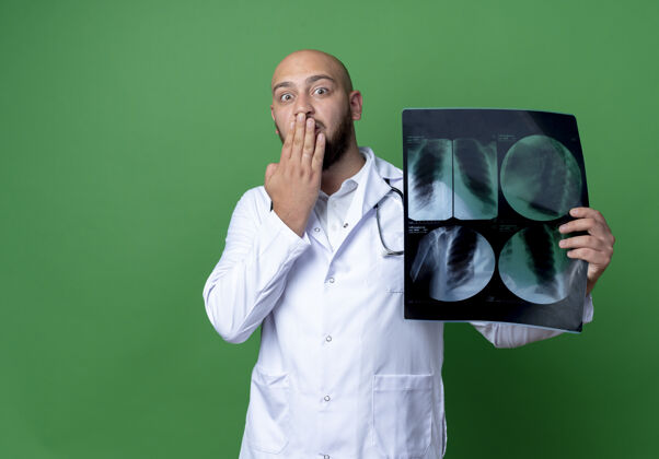 医疗年轻的男医生被吓坏了 穿着医用长袍 听诊器拿着x光片 手捂着嘴 绿色背景上被隔离听诊器医生X光