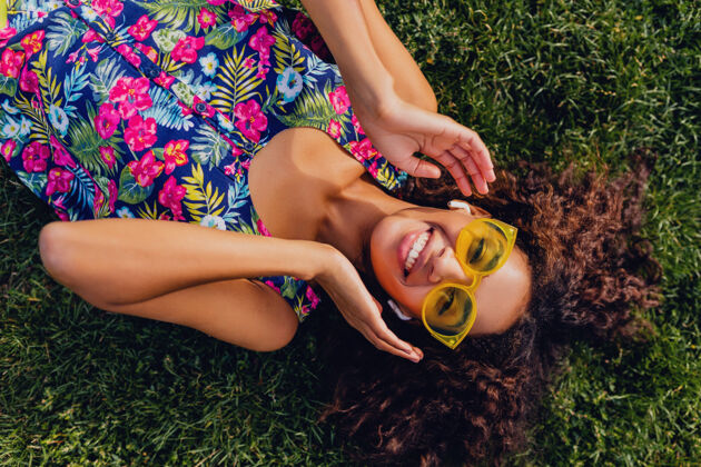 非洲裔美国人年轻时尚的黑人女子戴着无线耳机听音乐在公园里玩得很开心 夏日时尚风格 五颜六色的时髦装扮 躺在草地上 俯瞰风景积极肖像卷发