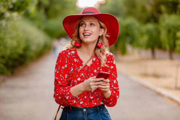 享受迷人的时尚金发微笑的女人在稻草红帽子和衬衫夏季时尚套装使用手机明亮脸女性