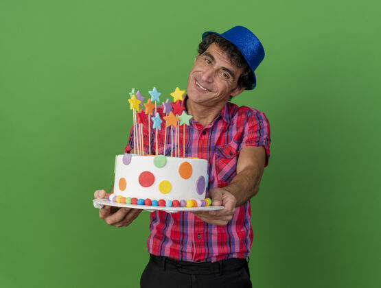 微笑微笑的中年白种人党男子戴着党帽子看着相机伸展生日蛋糕对相机隔离在绿色背景与复制空间蛋糕帽子空间