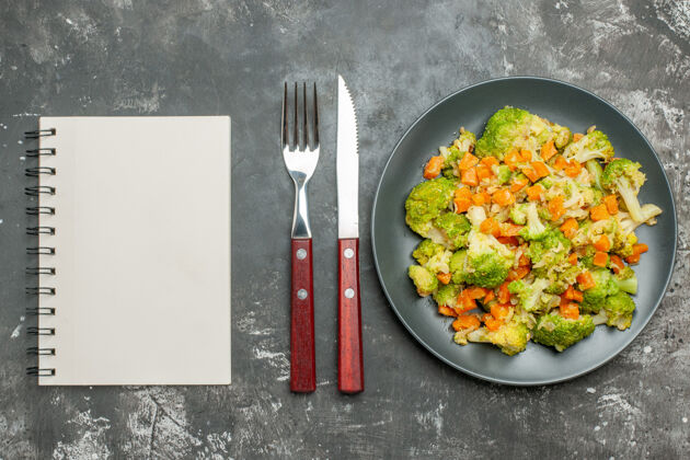 餐厅健康餐 西兰花和胡萝卜放在黑色盘子里 笔记本旁边放着刀叉笔记本餐具厨具