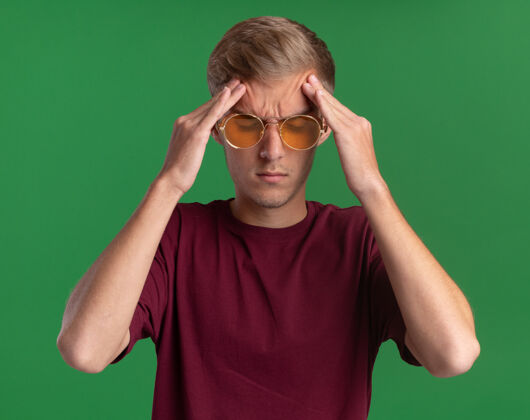 衬衫厌倦了闭着眼睛的年轻帅哥穿着红衬衫戴着眼镜把双手放在额头上隔离在绿色的墙上年轻疲倦眼镜