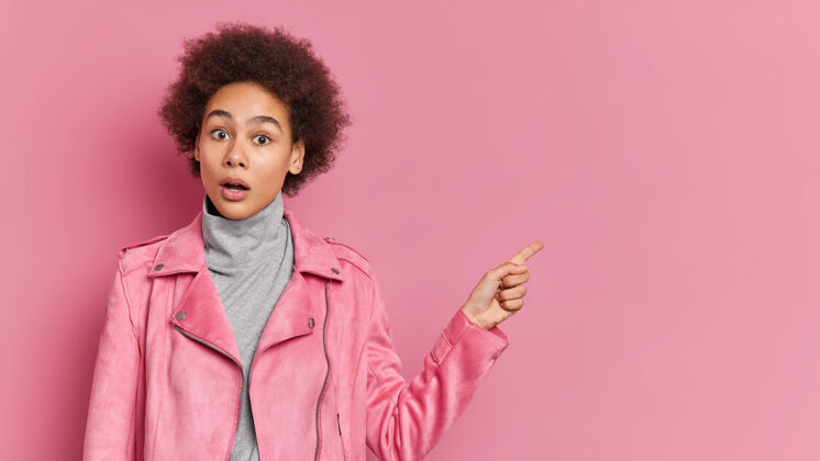 女性充满感情的非洲裔女青年 用手指着粉色墙上的复制空间 展示了一个惊人的宣传片惊奇惊艳肖像