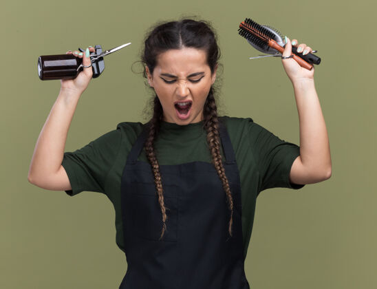 愤怒愤怒的年轻女理发师穿着制服举起理发工具隔离在橄榄绿的墙上制服理发师工具