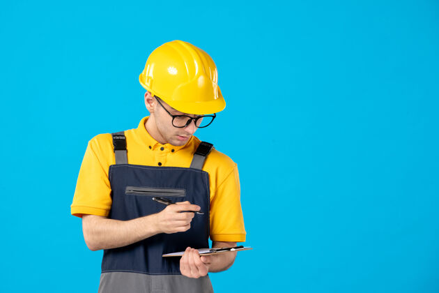 蓝色身穿制服和头盔的男建筑工人在蓝色墙上做笔记的正面图修理工制服工人