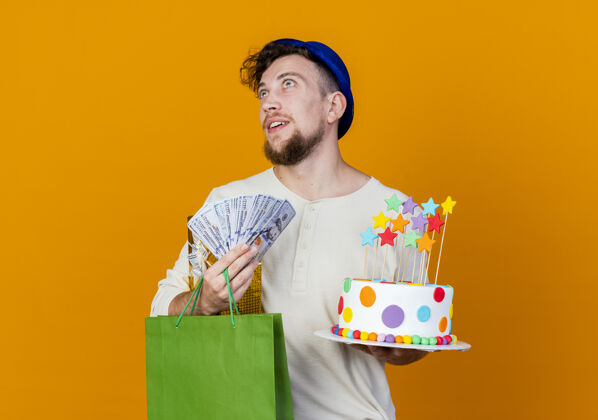 礼物令人印象深刻的年轻英俊的斯拉夫党人戴着党的帽子拿着礼品盒钱纸袋和生日蛋糕与星星仰望孤立的橙色背景与复制空间印象生日斯拉夫