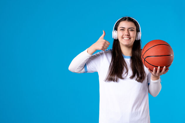 耳机正面图：戴着耳机拿着篮球的年轻女性站在蓝色的墙上成人年轻女性抱着
