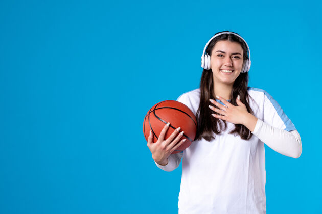 微笑正面图：戴着耳机拿着篮球的年轻女性站在蓝色的墙上年轻女性比赛蓝色