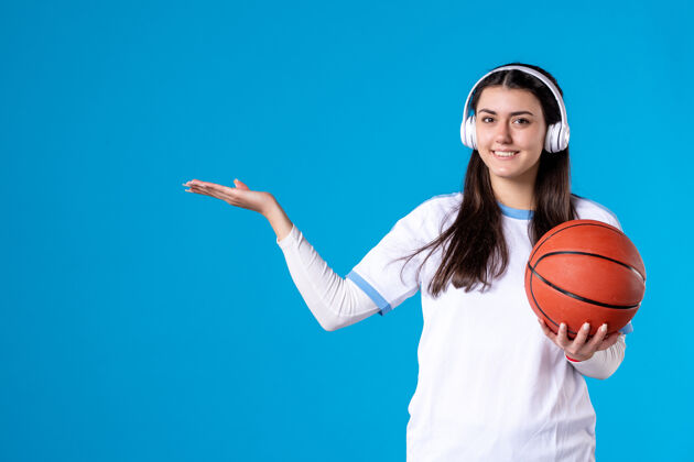 年轻女性正面图：戴着耳机拿着篮球的年轻女性站在蓝色的墙上手持成人微笑