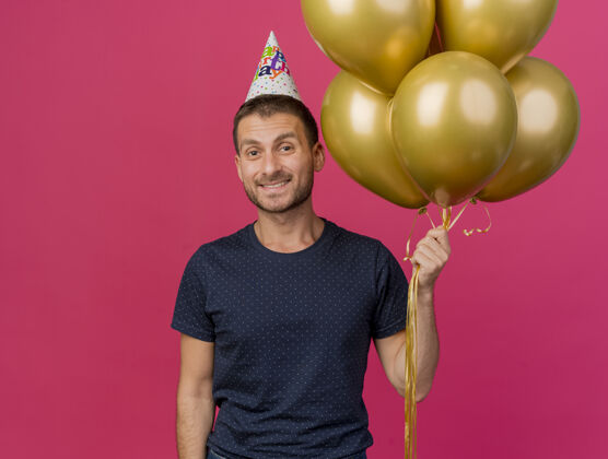 空间笑容可掬的高加索帅哥 戴着生日帽 手里拿着氦气球 背景是粉色的 还有复制空间复制气球帽子
