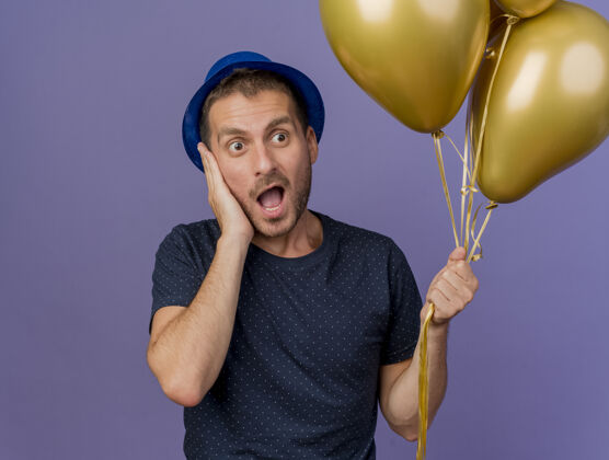 脸兴奋的白人帅哥戴着蓝色帽子把手放在脸上拿着氦气球隔离在紫色的背景和复制空间男人帽子紫色