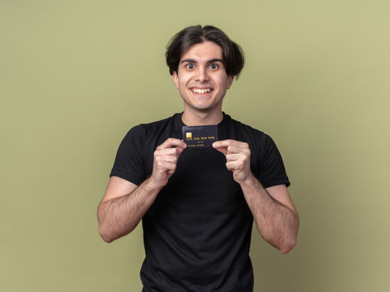 微笑微笑着的年轻帅哥穿着黑色t恤拿着信用卡隔离在橄榄绿的墙上信用卡T恤小伙子