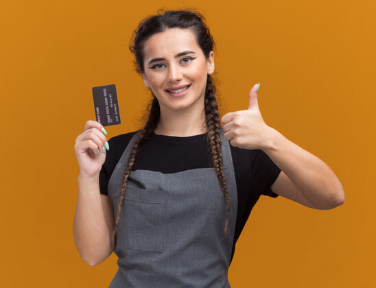 信用身着制服的年轻女理发师微笑着 手里拿着信用卡 竖起大拇指 孤零零地站在橙色的墙上理发师微笑大拇指