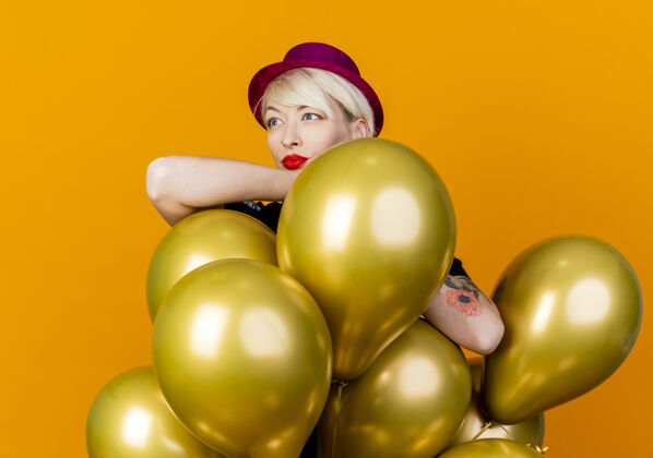 金发碧眼体贴的年轻金发派对女郎戴着派对帽站在气球后面 双臂搭在气球上 看着橙色背景下孤立的一面橙色派对女孩