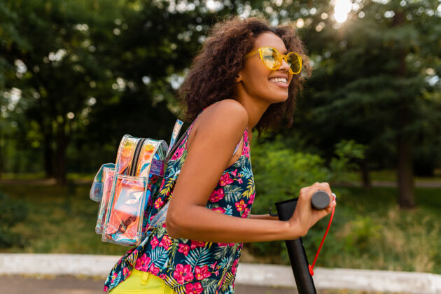 道路年轻时尚的黑人女子在公园里玩得很开心骑着电动脚踏车在夏天的时尚风格 五颜六色的时髦服装 穿着背包和黄色太阳镜积极摩托车欢呼