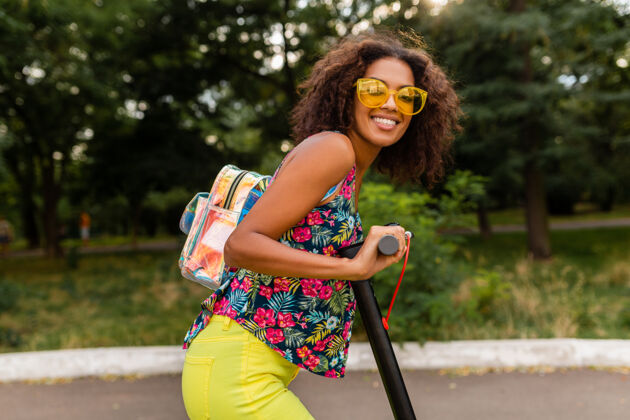 环保年轻时尚的黑人女子在公园里玩得很开心骑着电动脚踏车在夏天的时尚风格 五颜六色的时髦服装 穿着背包和黄色太阳镜太阳镜道路女孩